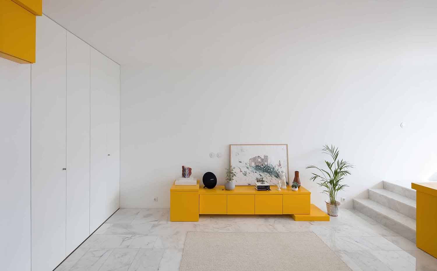 Mueble bajo amarillo sala de estar, armarios integrados, pavimento de mármol. Un mueble bajo de un intenso llamativo color focaliza la atención en la sala de estar donde se impone la austeridad. Junto a este, se oculta la zona de cocina tras paneles plegables que van de suelo a techo. 
