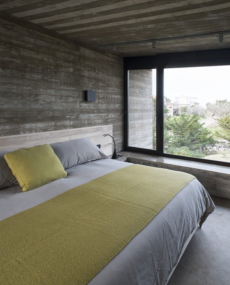 Dormitorio con paredes, suelo y techo de hormigón, gran ventanal al exterior, ropa de cama en blanco y verde pistacho
