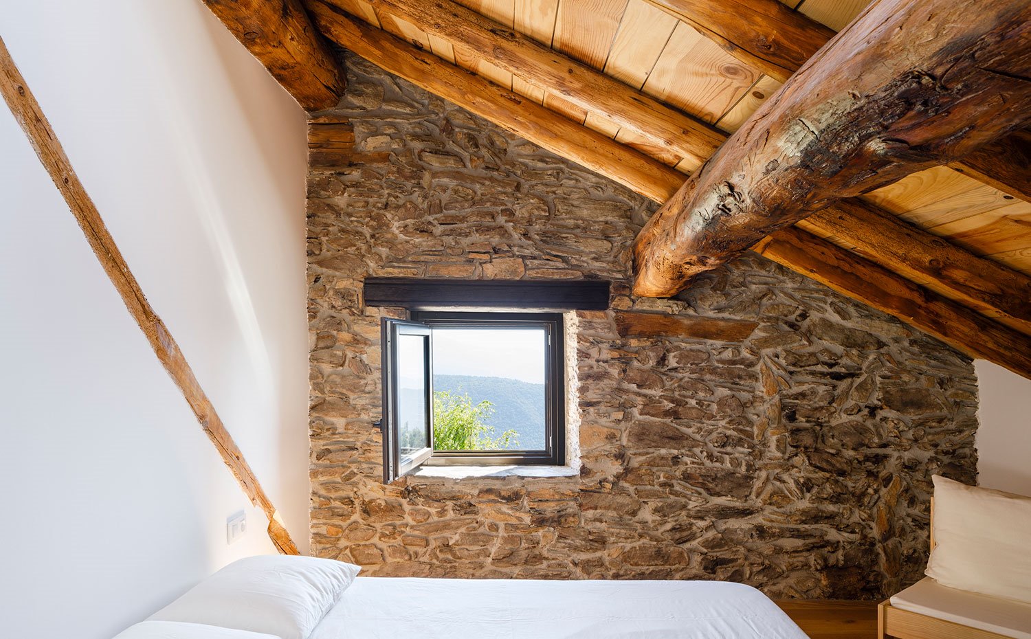 Detalle de ventana hacia el exterior con techo de madera en pendiente sobre cama de dormitorio