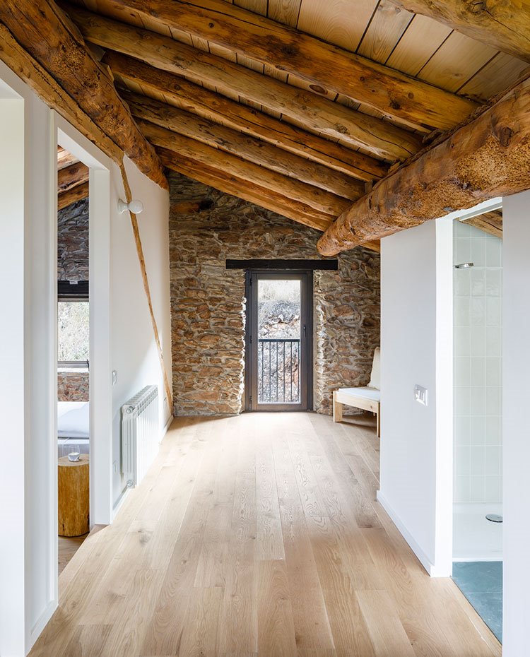 Zona de paso con suelo de madera, vigas en el techo y pared de fachada en piedra