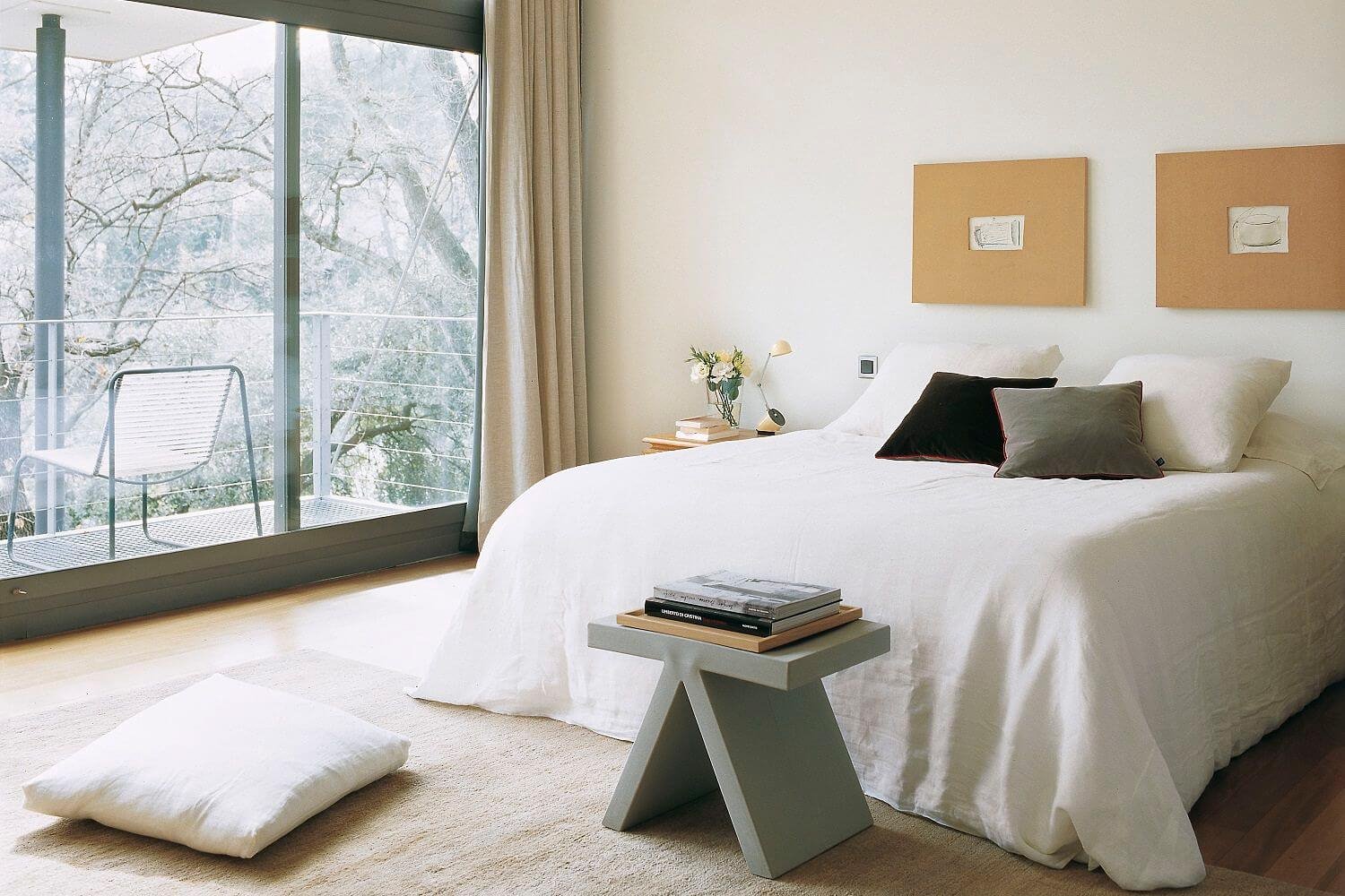 Dormitorio sin cabecero con cuadros en la pared y taburete a los pies de la cama. 