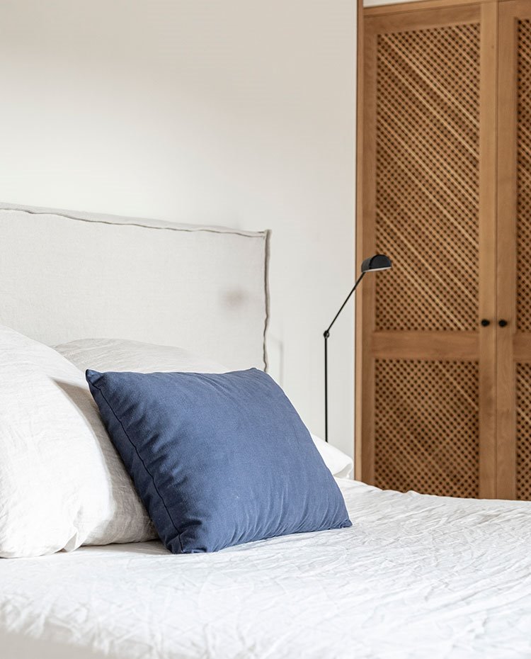 Cabecero de cama tapizado con tejido de lino, cojín azul, luminaria de sobremesa negra y armarios con celosía.