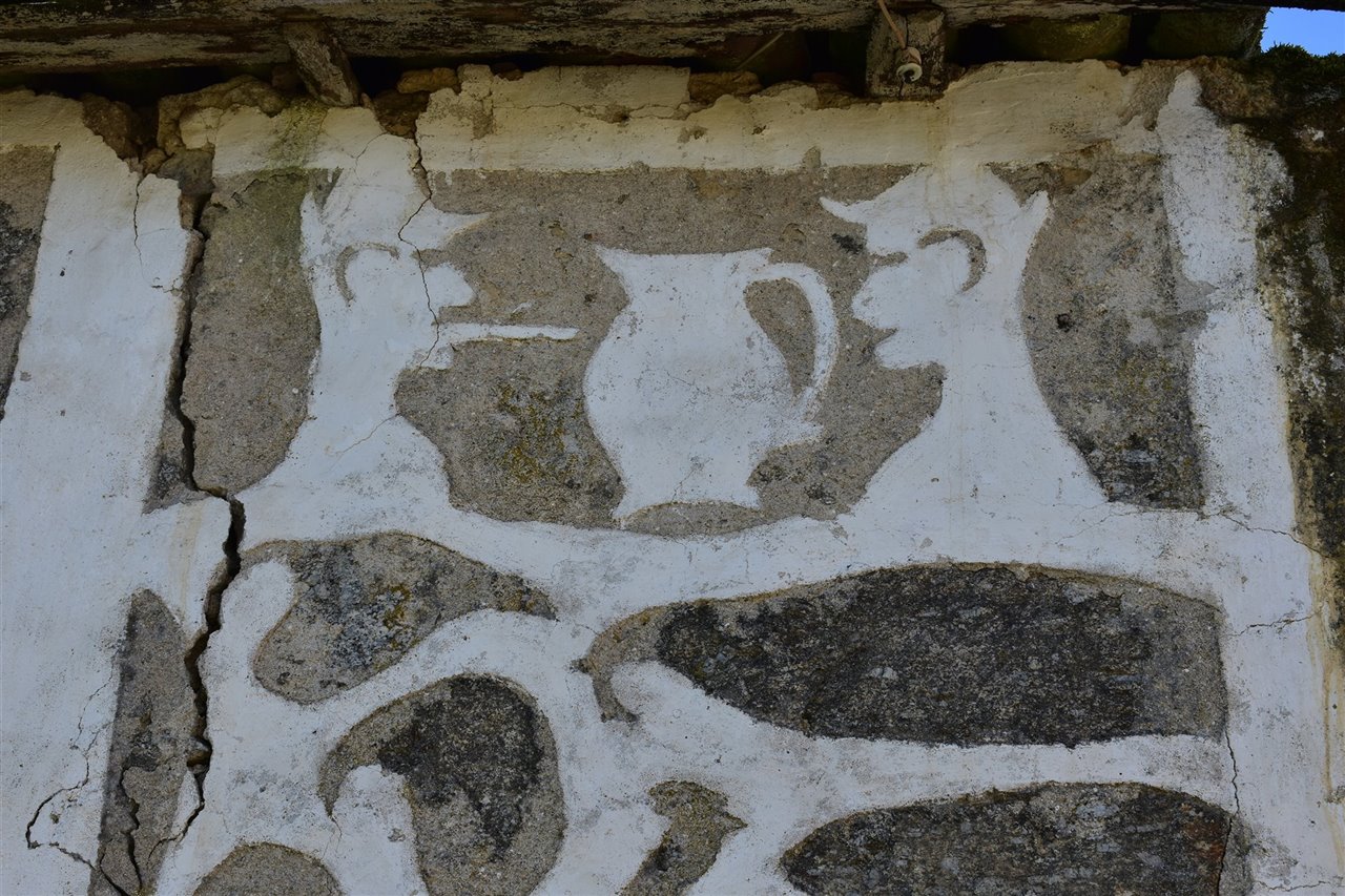 Detalle de la fachada anterior en el que se pueden apreciar dos bustos masculinos con sombrero y con cigarrillo y, en medio, un típico jarro de vino de cerámica de Gundivós, emblema de la Ribeira Sacra. 