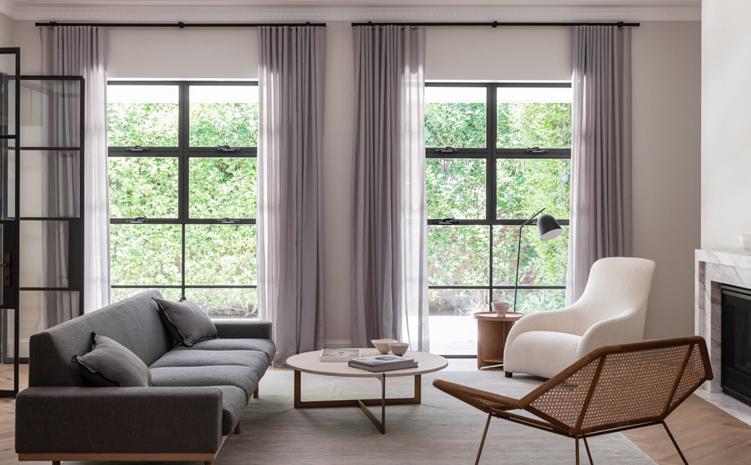 Salón con amplios ventanales provistos de cortinas grises, butaca cruda, sofá ris y asiento con estructura perforada, mesa de centro circular de madera