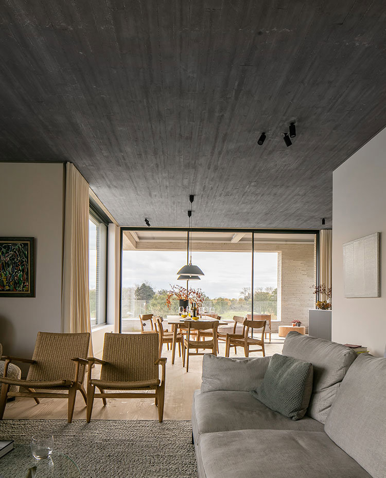 Salo´n con butacas de madera y fibras naturales, sofa´ en gris, techo de hormigo´n, al fondo mesa y sillas de comedor