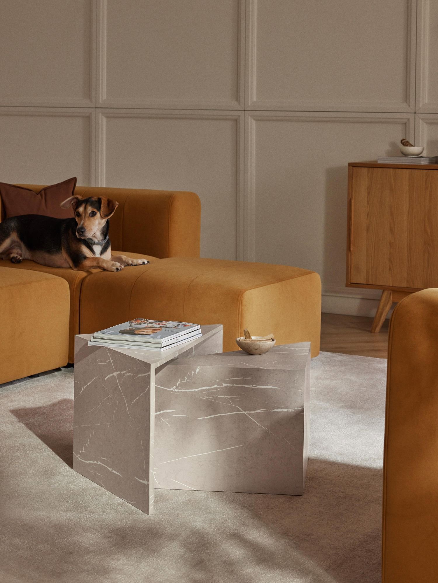 Mesa de marmol triangular con otra más pequeña en su interior en colores grises y blancos. Están en un saón moderno con sofá color mostaza, arriba de él un perro luce tranquilamente mirando a la cámara. 