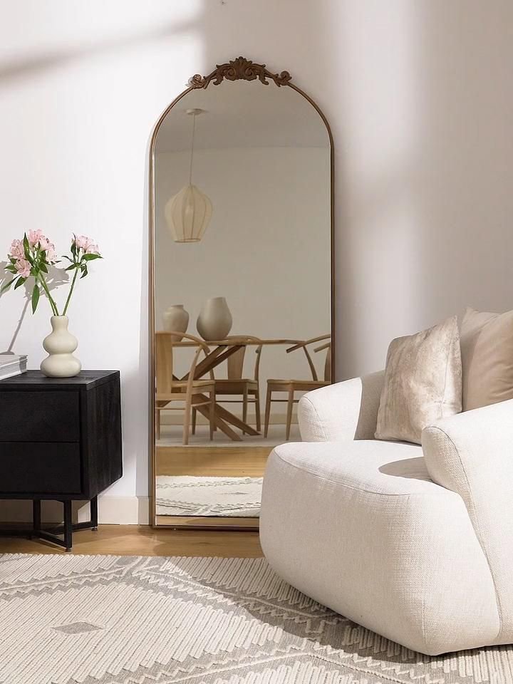Los espejos son parte de la decoración vertical como es el caso de este espejo grande y ovalado apoyado contra la pared del salón. Se ve un sillón mullido y profundo individual. Además, una consola negra adornada con un jarrón blanco con flores rosas. 