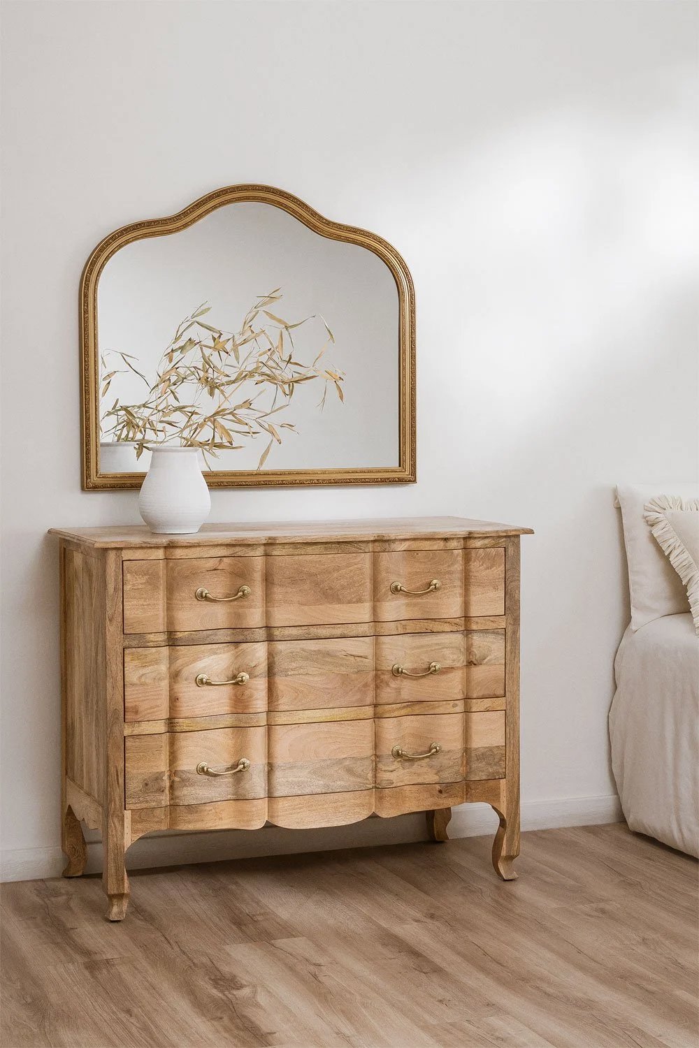 Cajonera cómoda de formas curvas como los muebles clásicos y elegantes. Madera clara que se complementa con el espejo de marco dorado sobre ella y el jarrón con flores y espigas. 