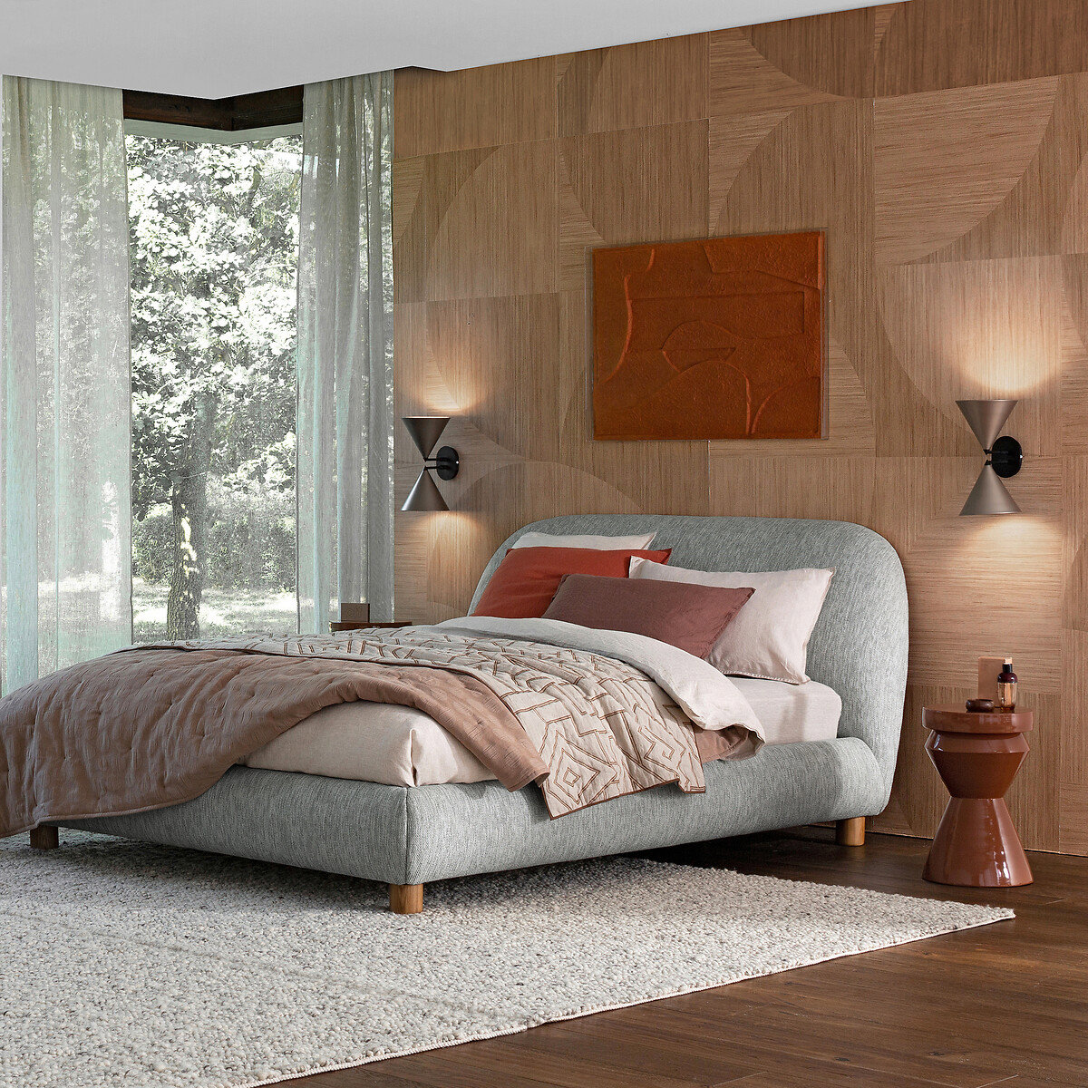 Habitación cálida de pared de madera y cuadro sobre el cabecero. La cama es robusta, en color gris y al costado un ventanal con luz natural. En el suelo se ha puesto una alfombra delgada en color gris. 