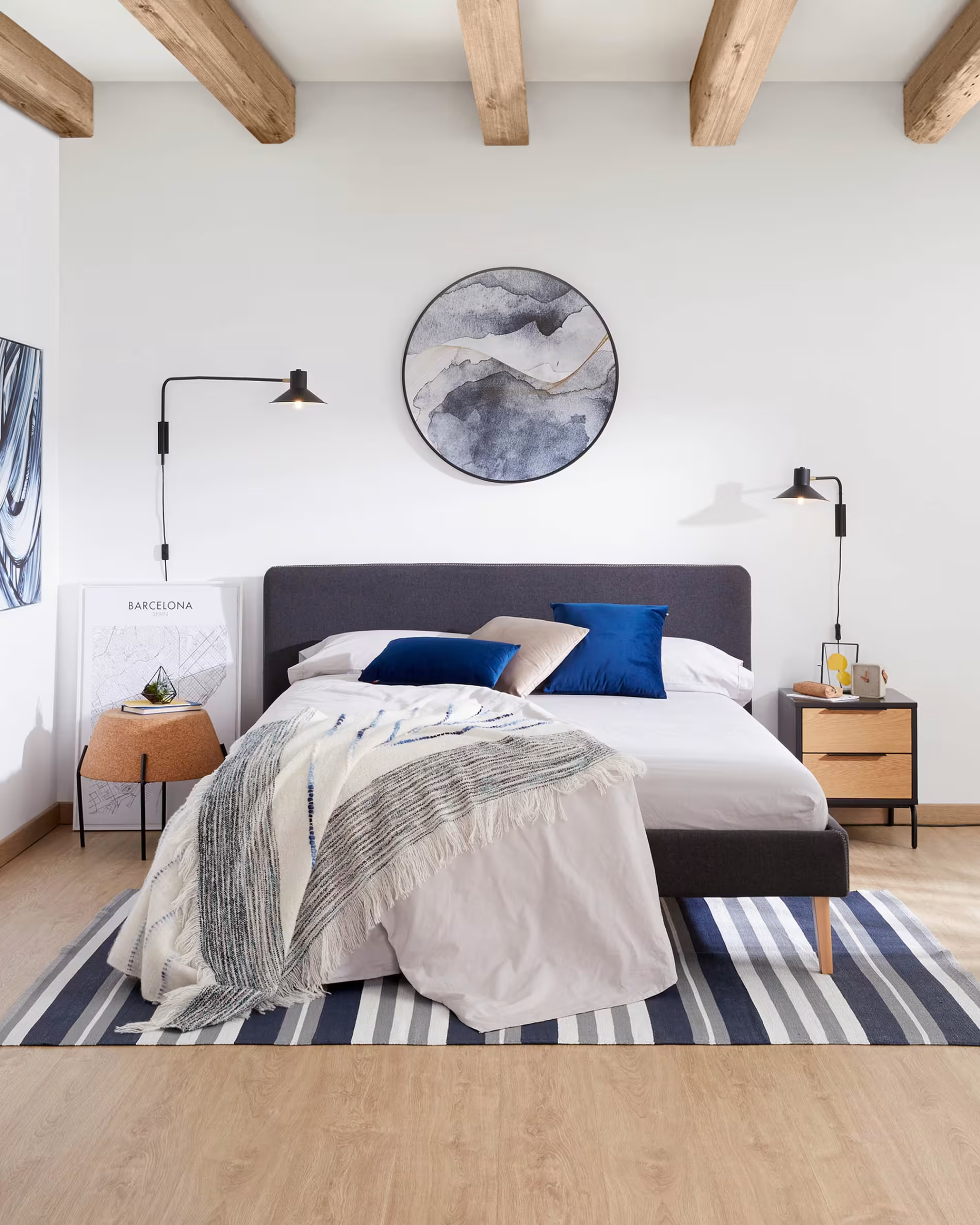 Cuadro redondo con pinturas en celeste y azul abstracto en habitación decorada de blanco, azul marino y gris con cama doble y techo con vigas a la vista. 