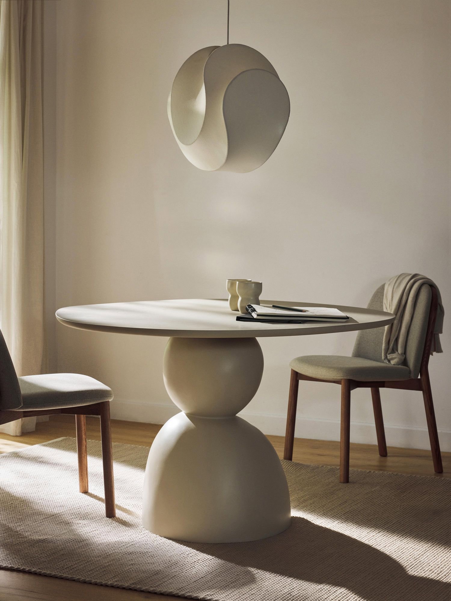 Comedor de estética sólida con dos bloques redondeados y una superficie redonda lisa. Sobre la mesa, una lámpara de diseños abstractos también en color blanco. 