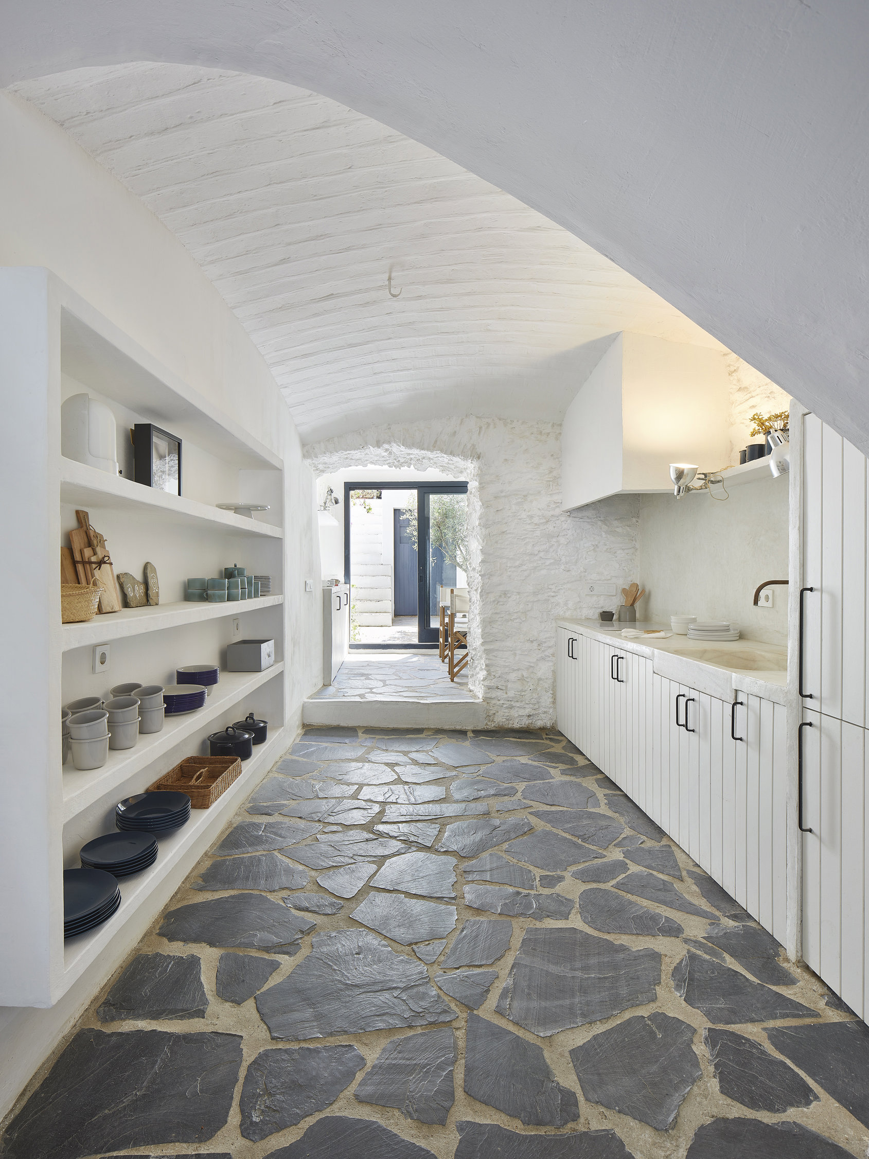 Cocina blanca con suelo de piedra en azul marino y vajilla expuesta en cerámica. Además, las paredes blancas y de piedra dan mucha luminosidad a esta cocina.