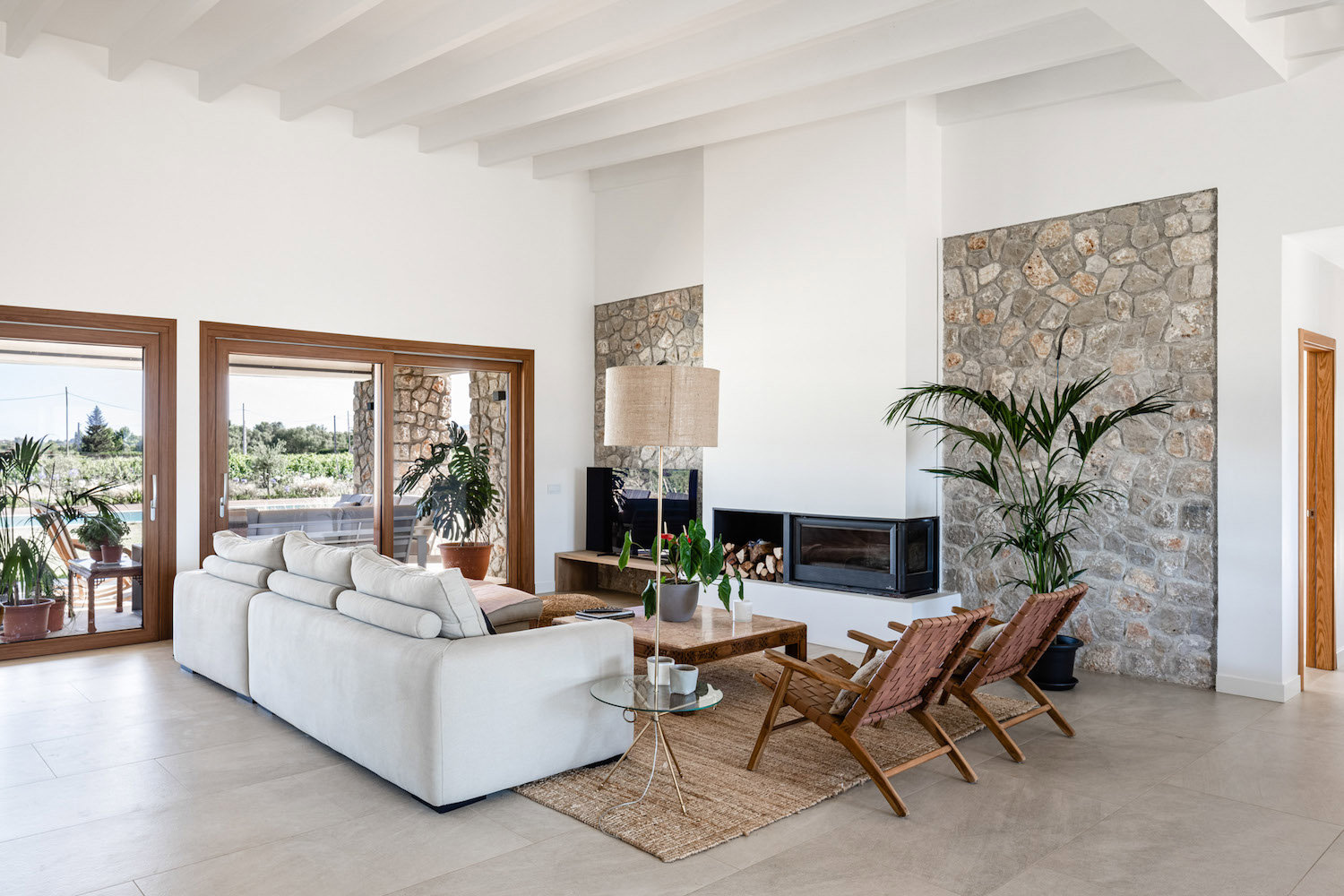 Salón diseñado de forma rústica y moderna con plantas, alfombra de yute, sofá blanco y elegante, chimenea elegante y moderna, pared de piedra y sillas en color terracota. 