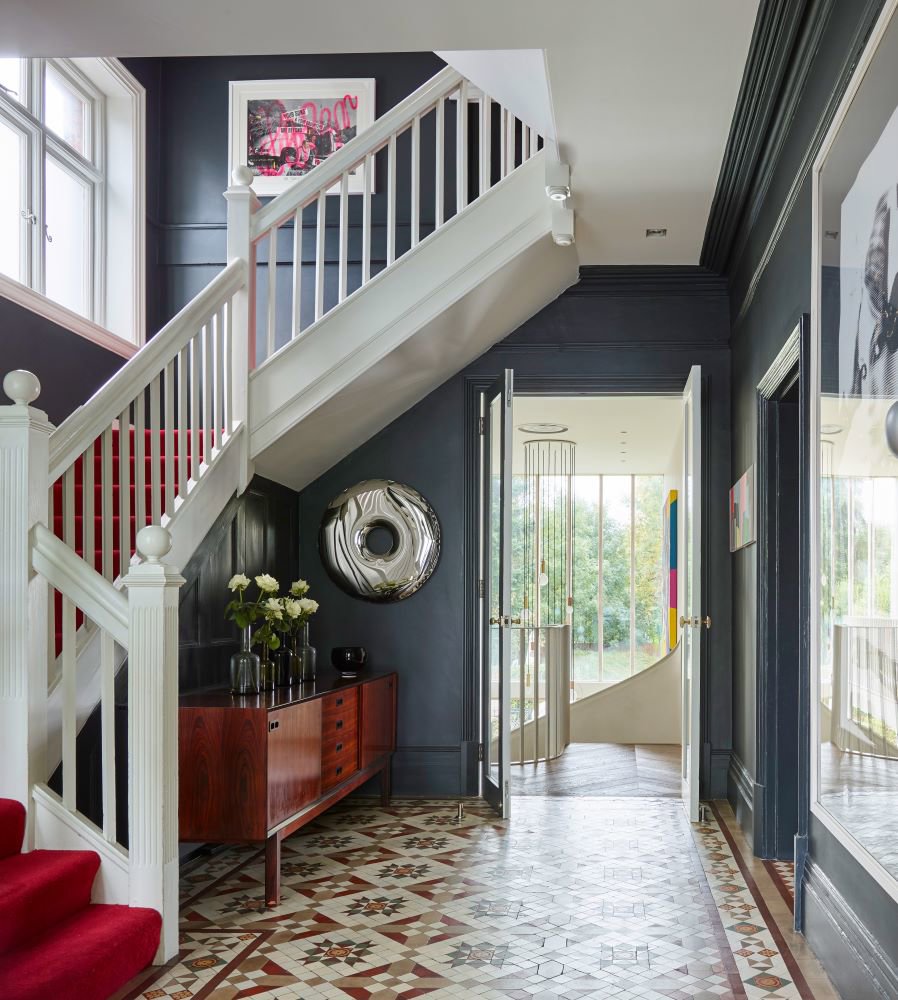 Recibidor amplio con escalera blanca recta y suelo de patrones geométricos, paredes azul piedra y entrada de luz natural. 