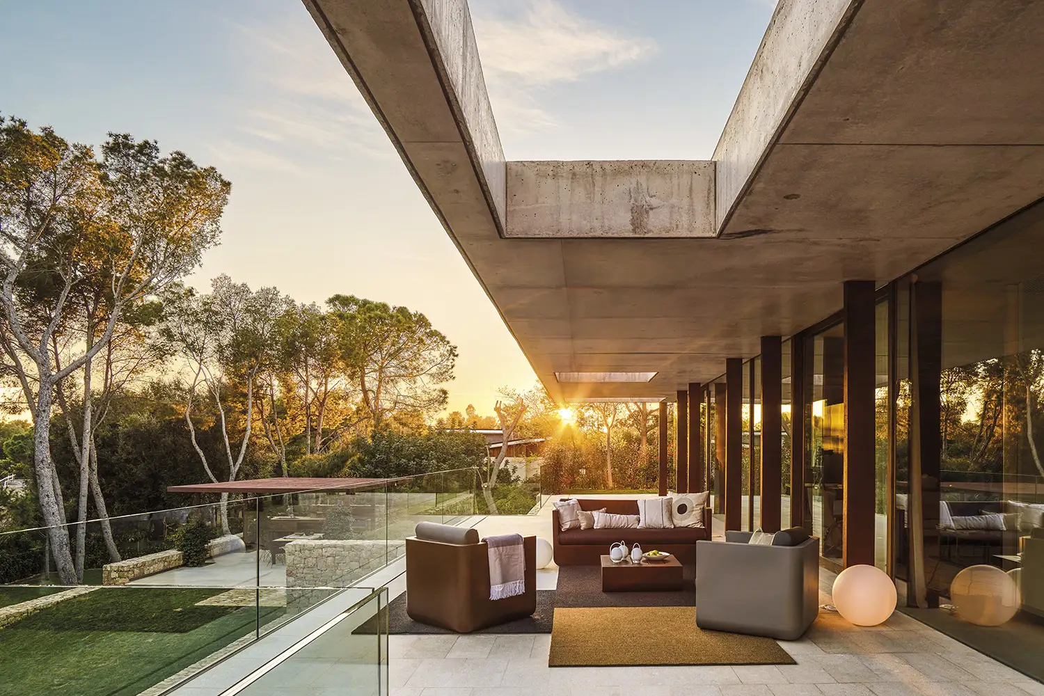 Terraza de balcón de cristal con modernos muebles en tonos marrones y grises y cuatro alfombras en el suelo. 