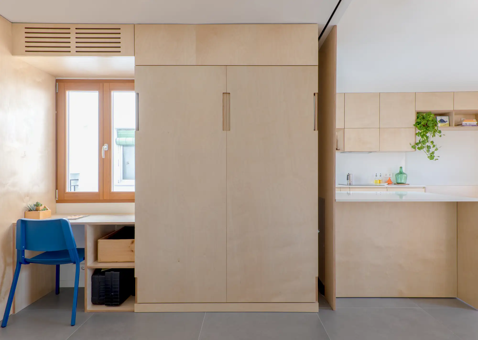 Habitación luminosa con madera clara, escritorio a la izquierda, silla azul eléctrico, armario de dos puertas con cama escondida dentro y cocina al costado derecho.