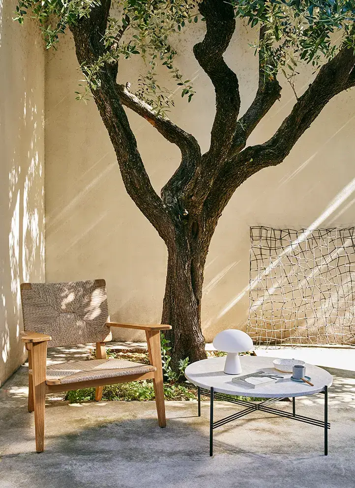 Jardín con árbol, silla de fibras naturales y estructura de madera acompañada de mesa de café de mármol y base metálica negra