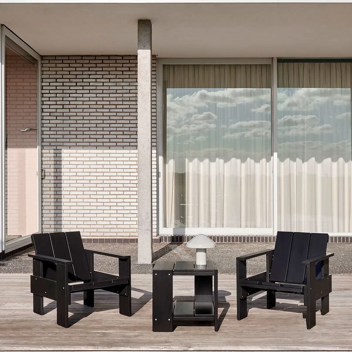 Conjunto de terraza de dos sillas y una mesita de madera negra en espacio iluminado con fondo gris y ventanal.