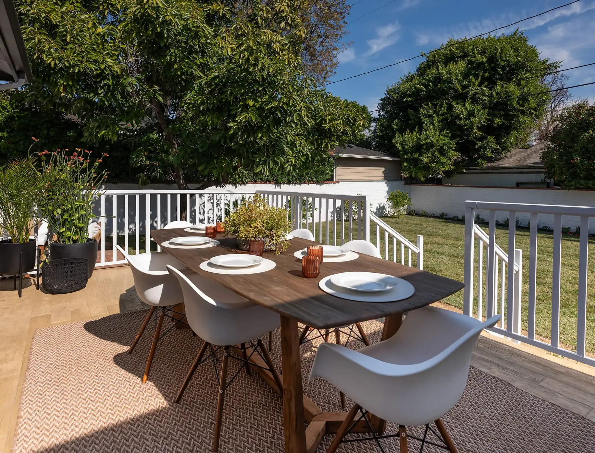 Comedor en terraza sobre alfombra de exterior, mesa rectangular de madera oscura y sillas blancas.