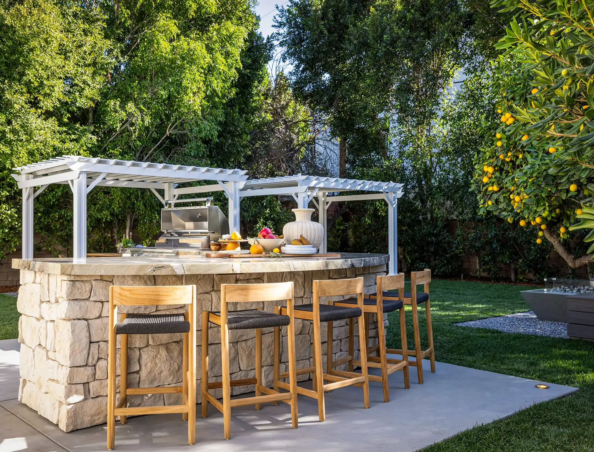 Cocina exterior diseñada por los hermanos Scott con encimera de piedra, sillas altas de madera y asiento gris y decoración blanca con pérgolas.