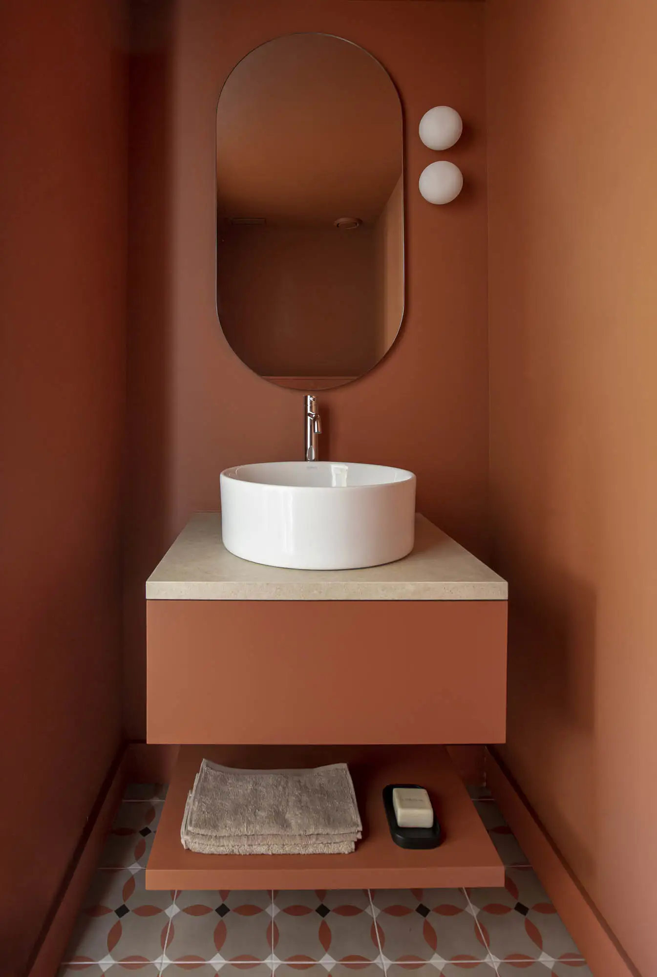 Baño pintado de color terracota, espejo ovalado, lavamanos blanco y superficie beige.