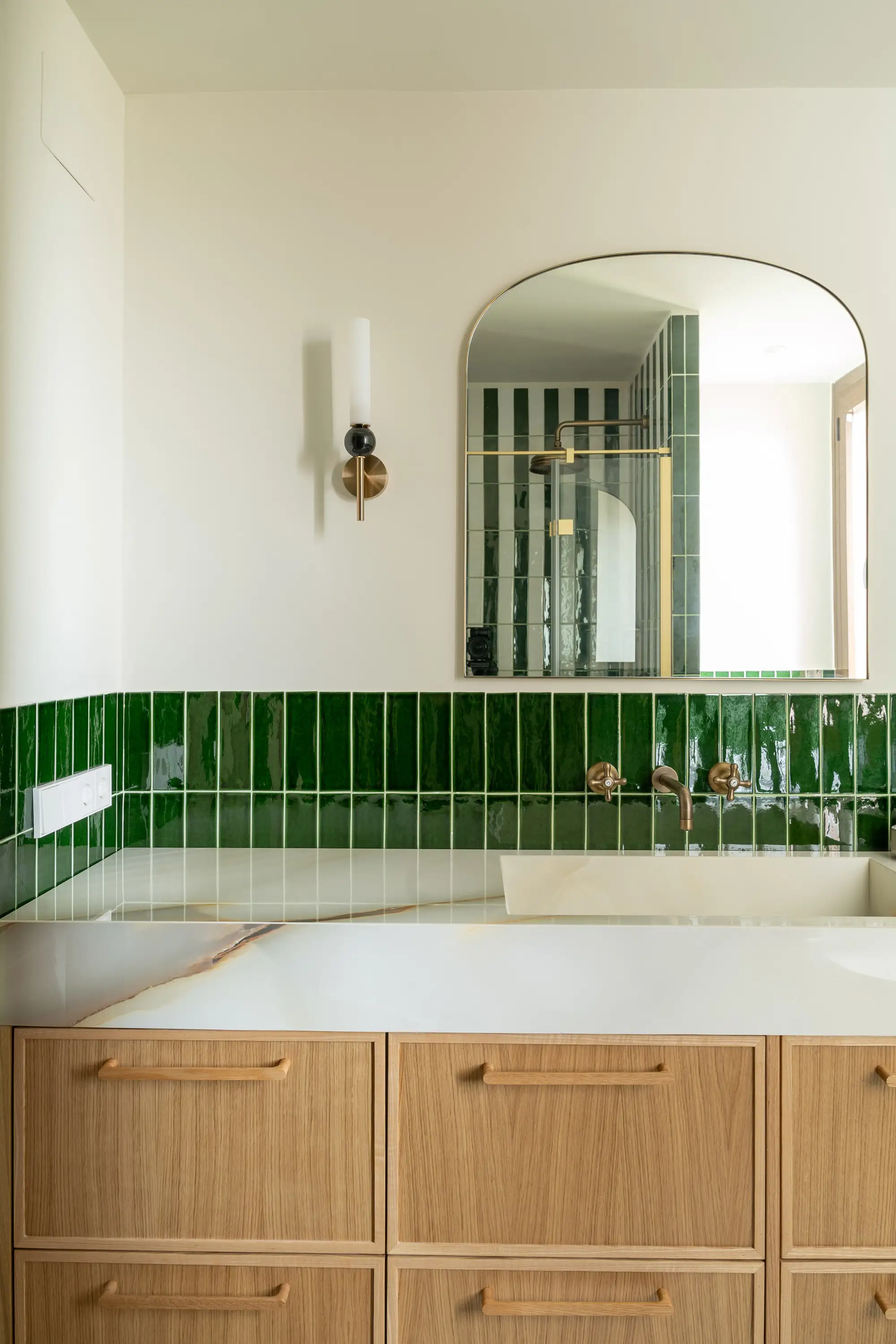 Baño con azulejos verdes, espejo curvado, cajones de madera y superficie de mármol.