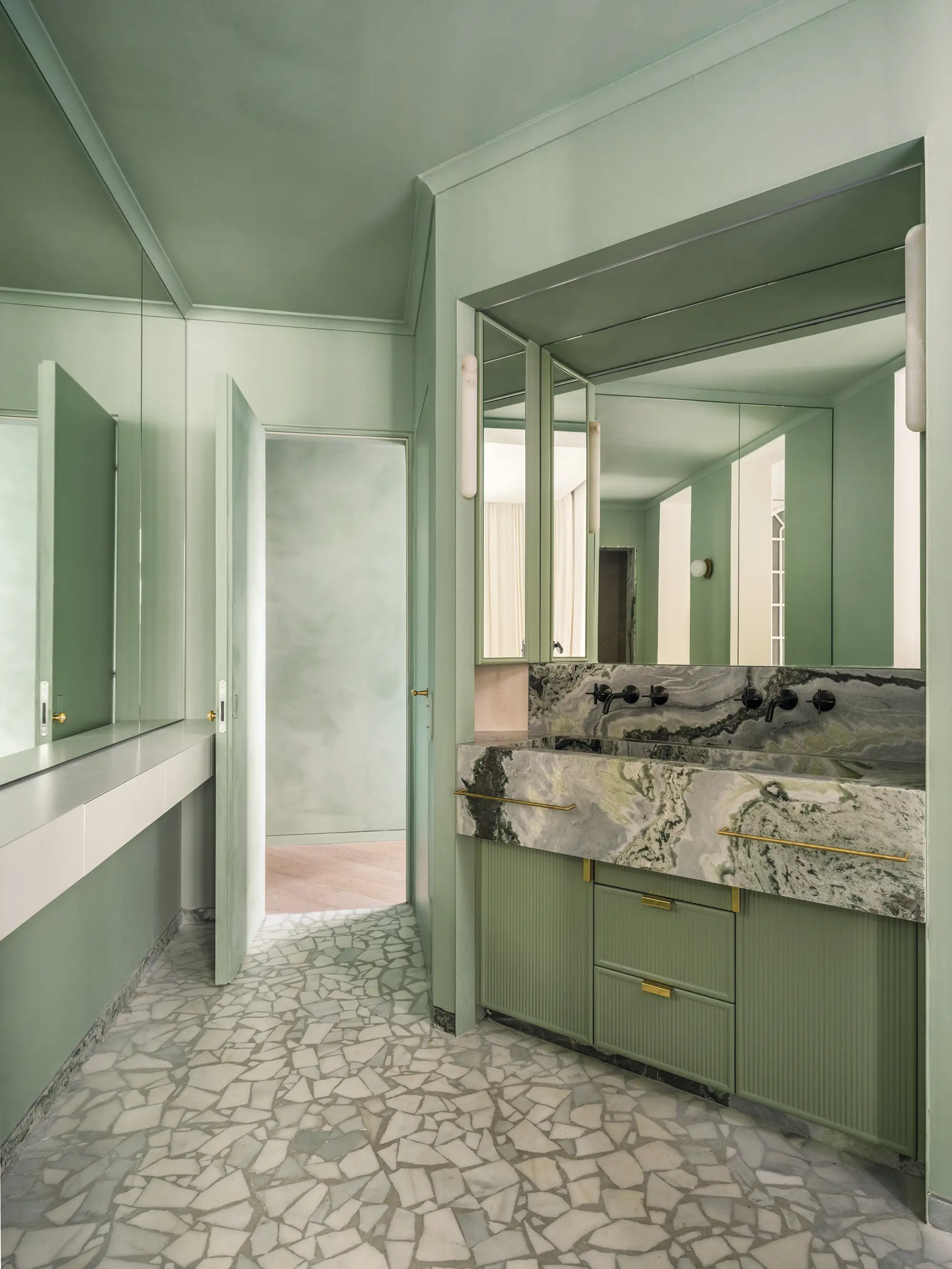 Baño pintado en color verde salvia, tono tendencia del 2024 y 2025. Suelo de piedra, encimera de mármol gris y tiradores dorados. Espejo grande rectangular.