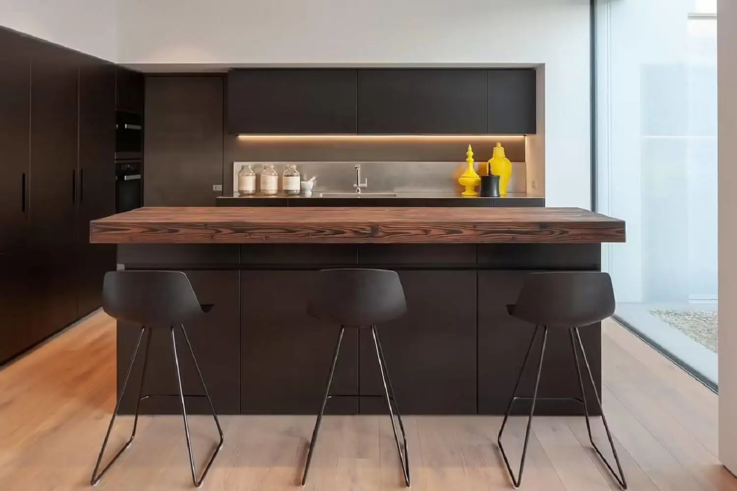 Cocina con mueble de color negro, encimera de madera con vetas negras y taburetes negros altos. 