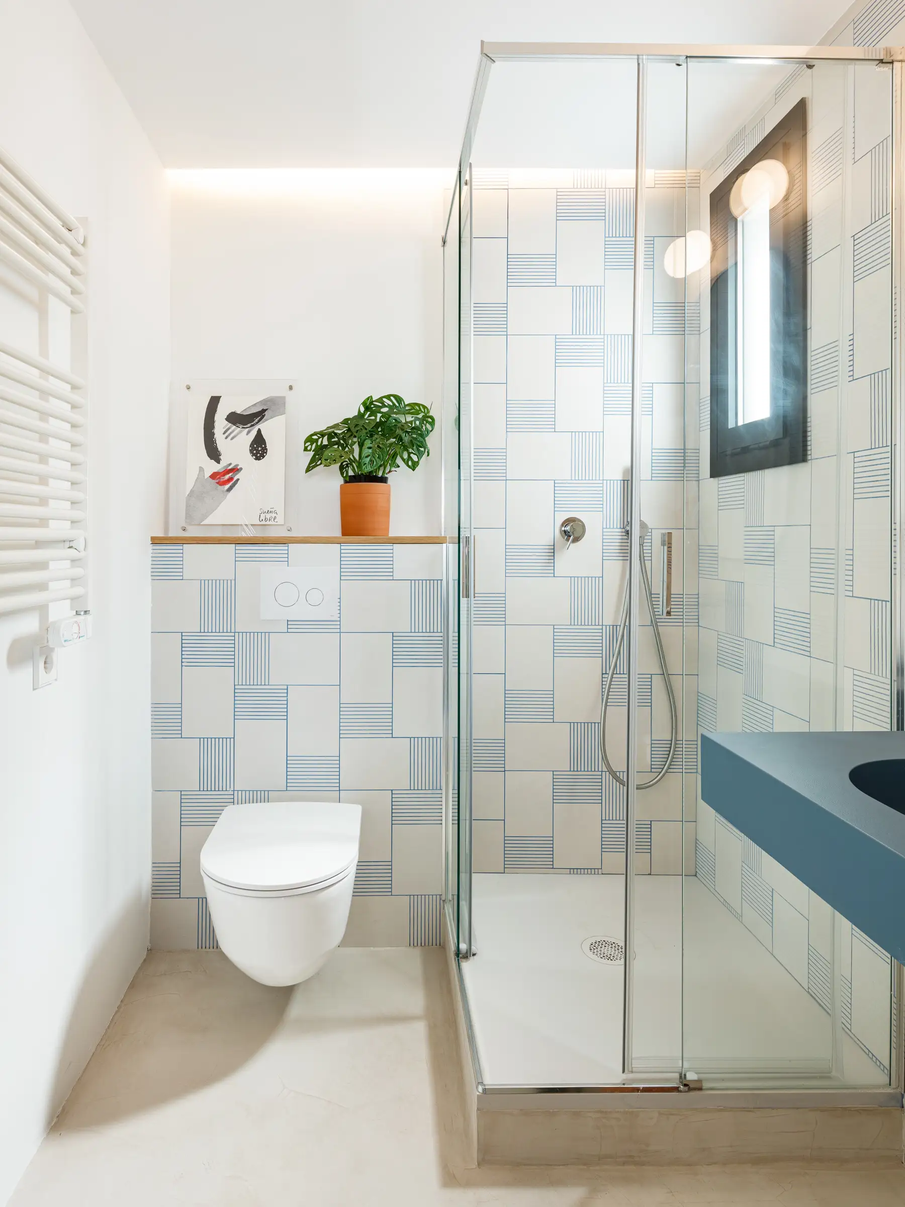 Baño en tonos celeste con encimera en azul cielo, inodoro blanco, azulejos blancos con rayas celeste, mampara de vidrio, planta pequeña y cuadro decorativo.