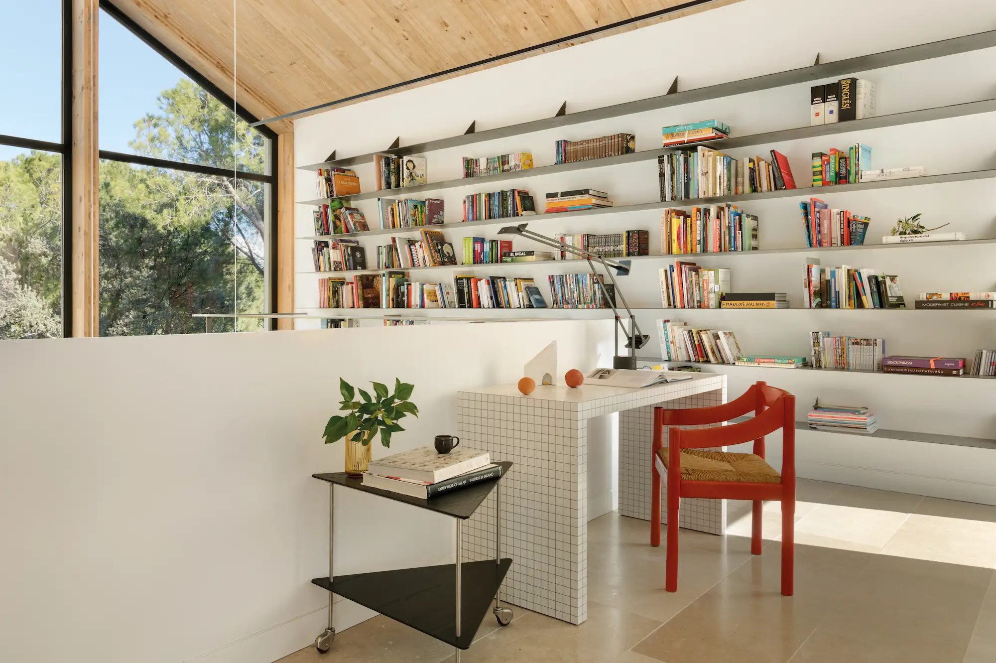 Estudio con gran estantería llena de libros, carrito negro con plana y escritorio blanco. 