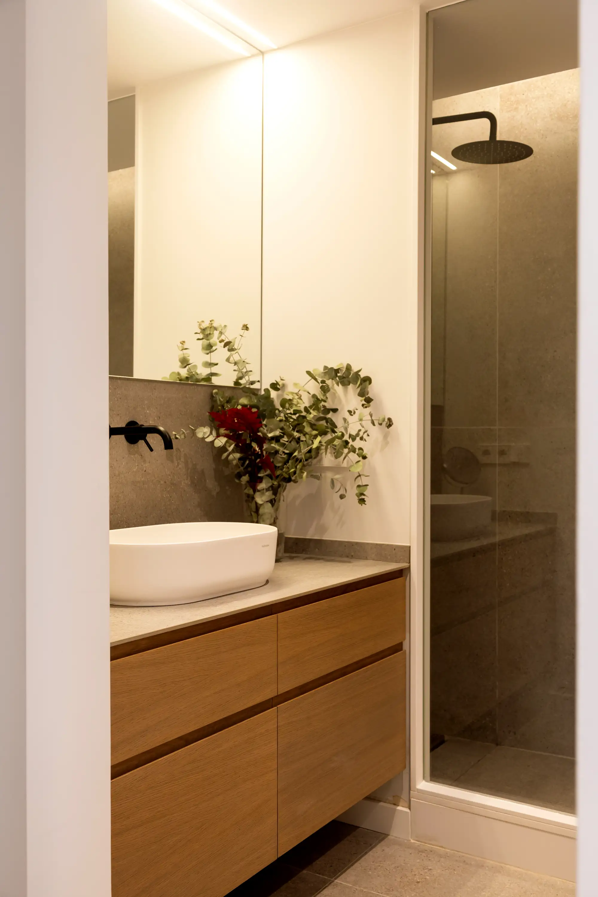 Baño con grifos negro, encimera blanca, mueble de madera, mampara fija con puerta de cristal a medida.