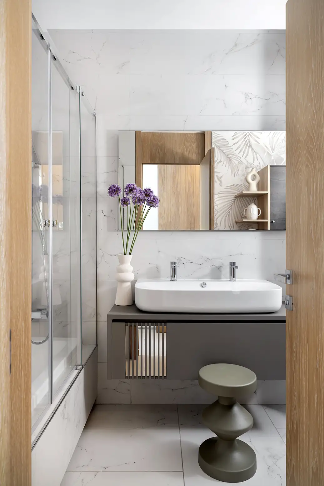 Baño con puertas de madera, mueble gris, taburete gris, flores violetas y mampara corredera de cristal.