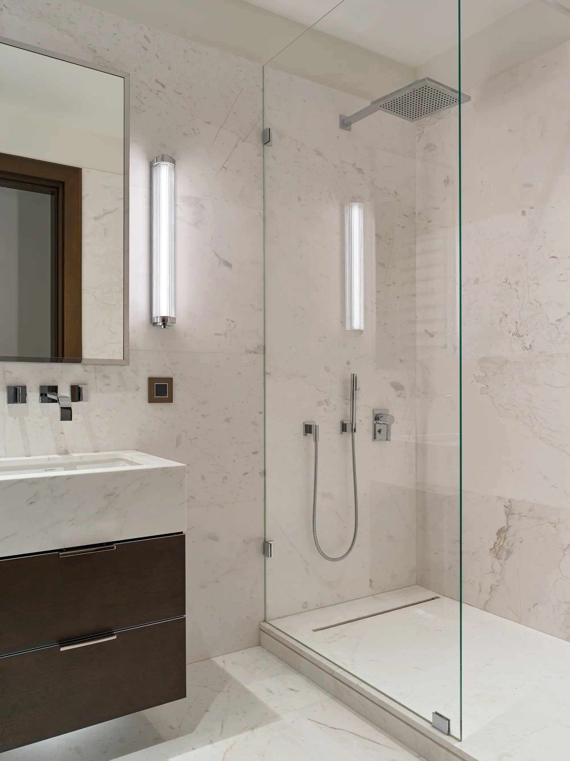 Baño con baldosas blancas con vetas naturales, encimera blanca, mueble de madera oscura, mampara de ducha de cristal de tipo fija. 