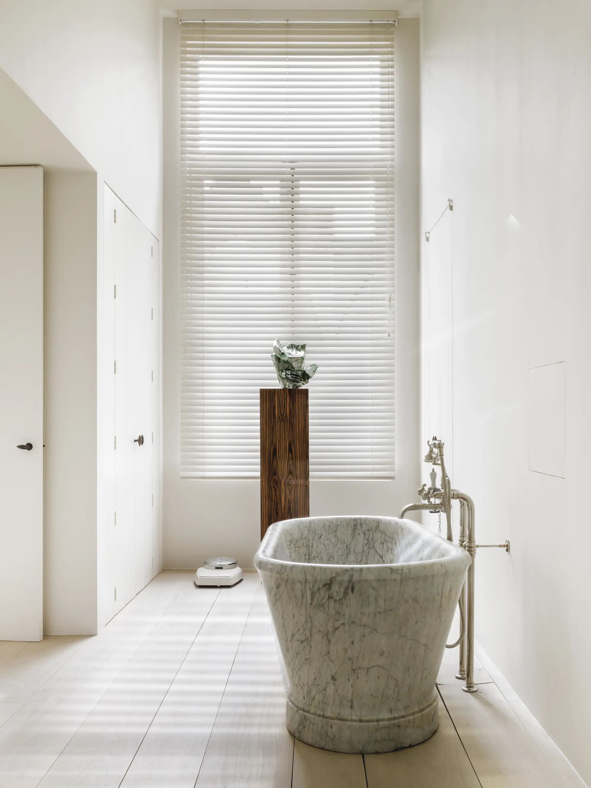 Baño luminoso con pilar de madera, escultura pequeña y bañera de mármol gris.