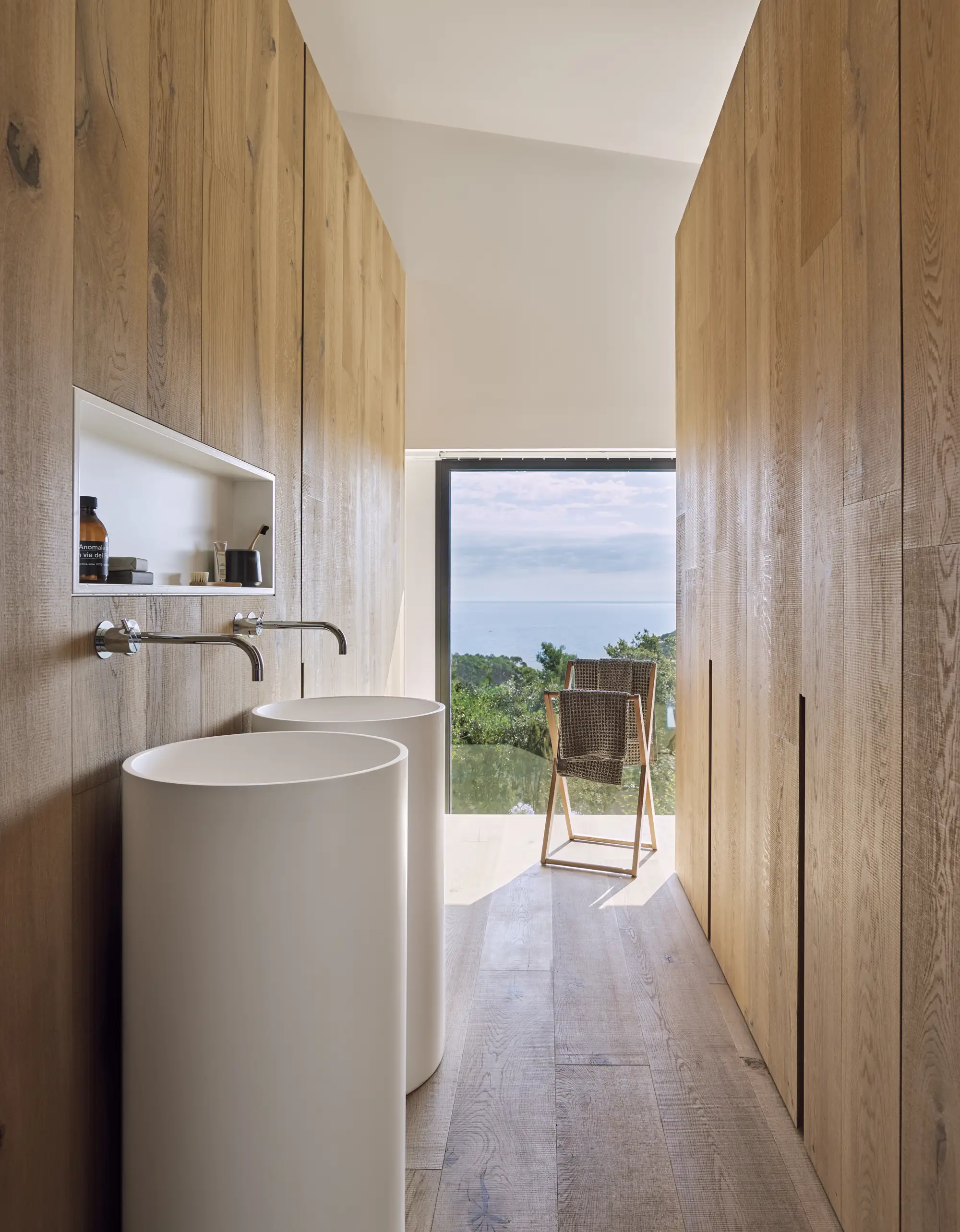 Baño con suelo de madera, revestimientos de madera, hornacina y doble lavabo con grifos cromados. Ventana al exterior con vistas.