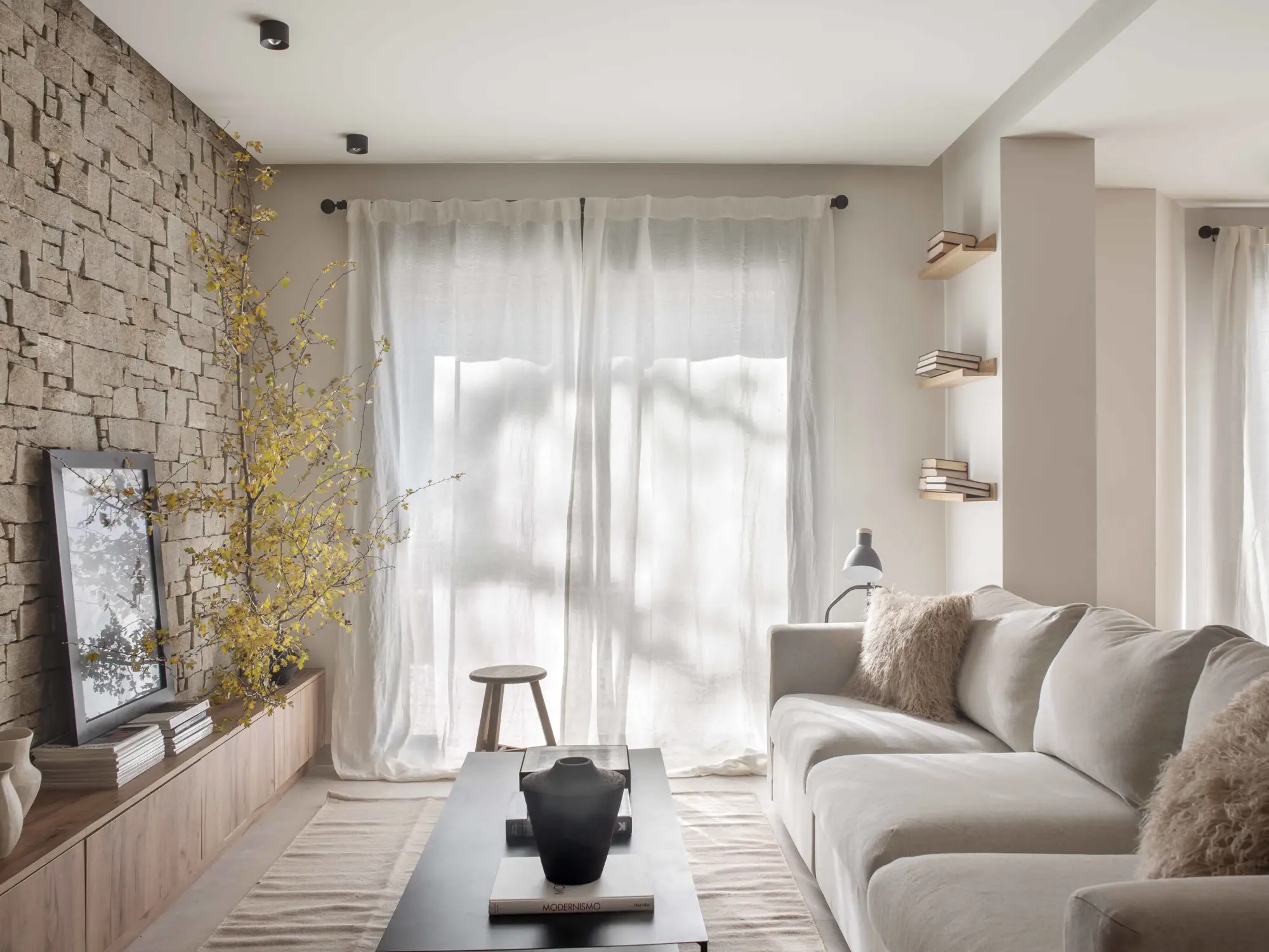 Salón pequeño de colores claros, pared de acento en piedra y muebles de madera. Baldas flotantes en una esquina.